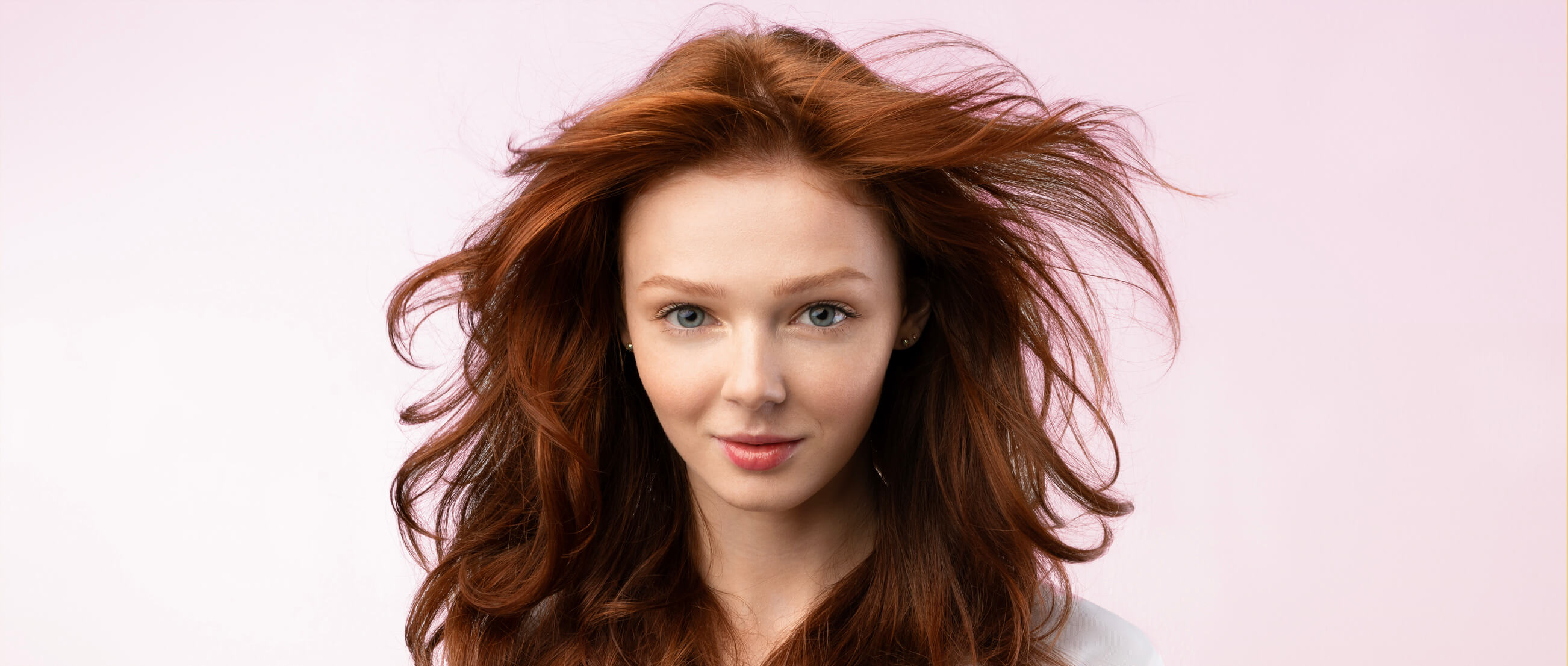 Jeune femme aux cheveux roux volumineux et éclatants, flottant librement, sur un fond rose doux.